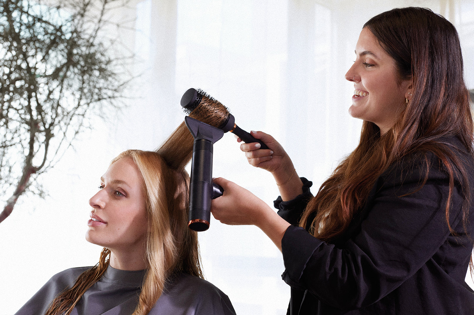 Woman styling another woman's hair with a hair dryer and brush outdoors.